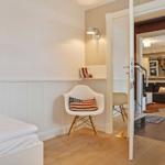 Schlafzimmer mit weißem Bett und Stuhl. Durchgang ins Wohnzimmer mit Sofa.