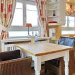 Dining room with table, chairs, and kitchen area. Windows with curtains and kitchen cabinets.