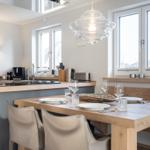 Dining area with wooden table, chairs, and windows. Kitchen in background.