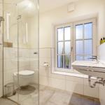Badezimmer mit Dusche, Toilette und Waschbecken. Fenster mit Pflanzen.
