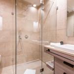 Bad mit Dusche, Waschbecken und Toilette. Glaswand, Holzschrank und Wandnische mit Blumen.