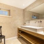 Badezimmer mit Waschtisch, Spiegel und Fenster. Holzregal und schwarze Stuhl.