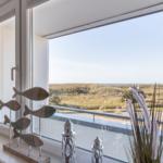 Blick auf Dünen und Meer durch große Fenster mit dekorativen Fischen und Pflanzen.