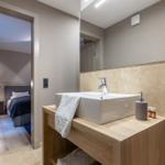 Badezimmer mit Waschbecken, Spiegel und Dusche. Schlafzimmer im Hintergrund sichtbar.