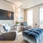 Wohnzimmer mit Sofa, Sessel und blauem Sitzkissen. Große Fenster und Fernseher an der Wand.