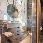 Badezimmer mit Dusche, Waschbecken und Toilette. Dunkle Fliesen und Holzmöbel.