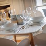 Tisch mit weißen Tellern, Gläsern und Besteck. Weiße Stühle und Fenster im Hintergrund.