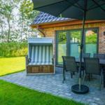 Terrasse mit Gartenliege, Tisch und Stühlen unter Sonnenschirm. Hintergrund: Grasfläche und Bäume.
