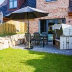 Terrasse mit Tisch, Stühlen und Sonnenschirm neben Haus. Weiße Strandkorb neben Steinmauer.