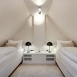 Zwei Betten mit weißen Laken und Kissen, schwarze Nachttische mit Lampen, schräges Dach und weißer Wand.
