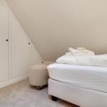 Schlafzimmer mit weißem Bett, Kleider schrank und Sitzhocker unter schräger Decke.