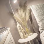 Bettkopfteil mit Kissen, Nachttisch mit Vasen und Pampasgras, Stehlampe und Fensterbank.