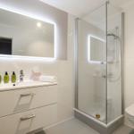 Badezimmer mit Dusche, Waschbecken und Toilette. Mit beleuchtetem Spiegel und grünen Flaschen.