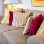 Ein Sofa mit mehreren Kissen in Rot und Gelb. Eine Lampe und ein Bild sind im Hintergrund sichtbar.