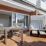 Deck with wooden table, chairs, and beach chair. Background: living area with window.