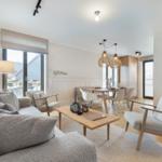 Living and dining area with sofa, chairs, and table. Wooden and metal furniture. Windows and ceiling lights.