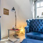 A room with blue armchair, windows and table. Wall art and lamps are visible.