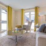 Wohnzimmer mit Sofa, Sessel, Glas-Tisch und gelben Vorhängen.