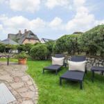 Backyard with loungers, table, and green areas. Stone wall and trees in the background.