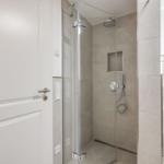 Shower cabin with glass enclosure, showerhead, and handheld shower. White door and vanity with mirror.