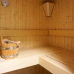 Holzsauna mit Sitzbänken und Eimer. Wandthermometer und Holzverkleidung sichtbar.