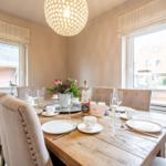 Dining table with chairs, flowers, and wine bottle by windows overlooking houses.
