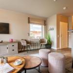 Living area with TV, seating, and window. Wooden table, plants, and bar stools visible.