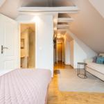 Schlafzimmer mit Bett und Sofa unter Dachschrägen. Blick in Badezimmer und Flur.