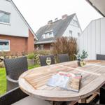 Terrasse mit Holztisch, Stühlen und Wein. Hintergrund: Haus und Garten.