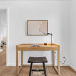 Schreibtisch mit Stuhl, Lampe und Bild an der Wand. Durchgang zu Schlafzimmer.