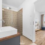Badezimmer mit Dusche und Waschbecken. Nebenraum mit Bett und Fenster.