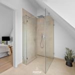 Shower cabin with glass enclosure and showerhead. Bedroom with bed and window in background.