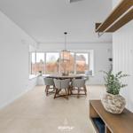Dining room with round table, four chairs, and large window. Wooden shelf with plant on right.