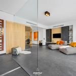 Modern living area with sofa, TV, and glass wall. Wooden doors and plants visible.