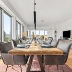 Dining area with long wooden table and chairs, large windows and TV.