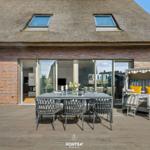 Terrace with dining area and beach chair in front of a house with thatched roof and glass facade.