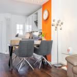 Dining area with table, chairs, and kitchen. Orange wall, lighting, and sofa visible.