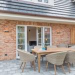 Terrace with wooden table and chairs in front of brick house with glass door.