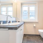 Bathroom with sink, mirror, and toilet. Window with view of rooftops.