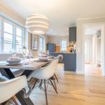 Dining area with table, chairs, and windows. Kitchen in background.