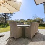 Terrasse mit Tisch, Stühlen und Sonnenschirm. Hintergrund: Gras, Bäume und Hauswand.