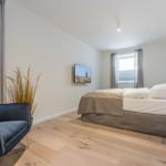 Schlafzimmer mit Bett, TV und Sessel. Holzboden und Fenster mit Vorhängen.