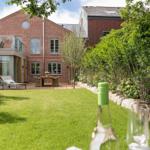 Red brick house with garden, terrace, and green lawn. Foreground: wine bottle and glass.