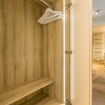 Closet with wooden walls and hangers. View into bedroom with bed and nightstand.