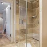 Shower with glass enclosure and stone tiles. View into hallway with wooden floor.