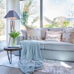 A sofa with cushions and blanket in front of large windows. Table with flowers and lamp beside it.