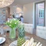 Dining table with chairs, flowers, and fruit. Kitchen and hallway in background.