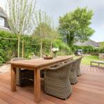 Wooden table with chairs on terrace. Green areas and trees in background.