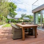 Wooden table with chairs on terrace beside modern house with glass facade.