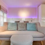 A sofa with cushions in front of a wall with LED lighting and a window.